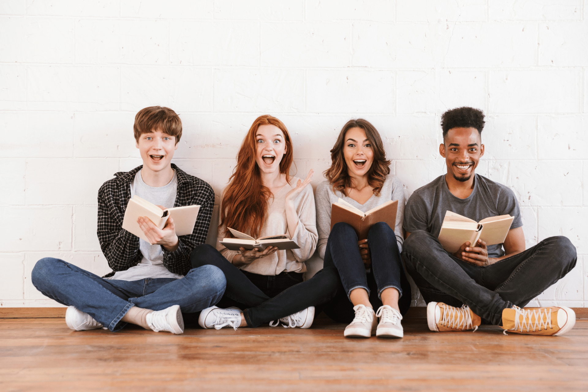 Four smiling young adults sitting on the floor and holding open books